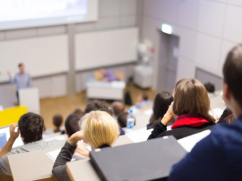 Professor giving presentation in lecture hall at university. Participants listening to lecture and making notes.