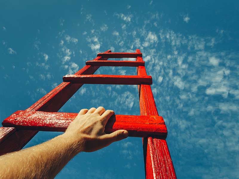 A male hand holds onto the crossbar of a red wooden staircase leading to the blue sky