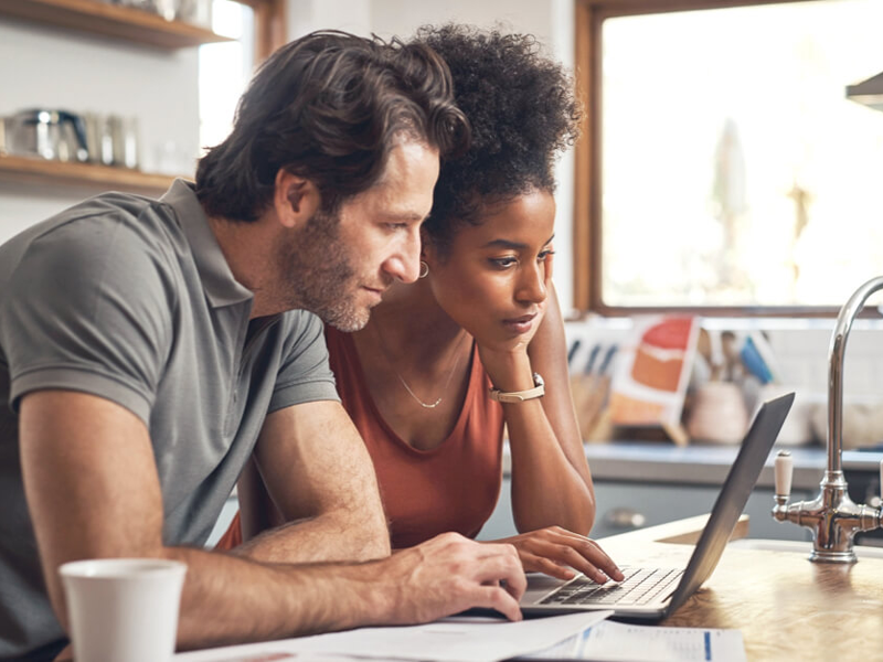 Man and woman look at a computer screen and documents in a kitchen.||