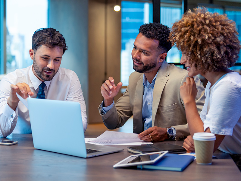 Three people in formal business wear meeting and looking at a laptop in workplace setting.