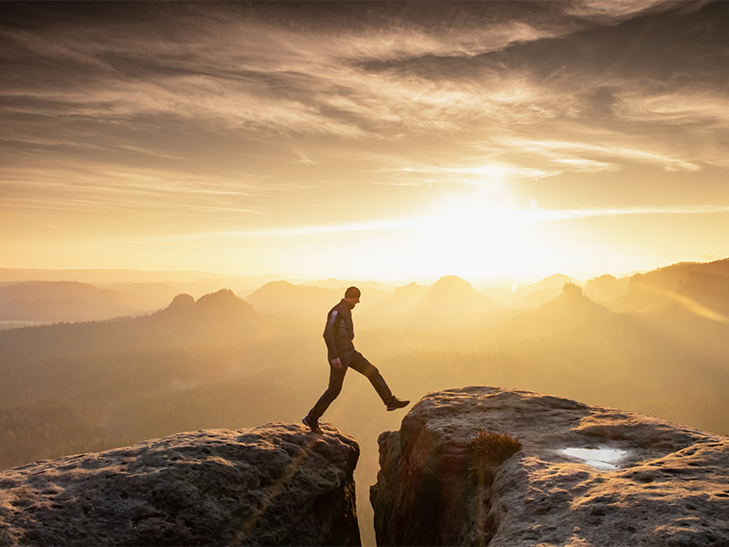 Man in hiking outfit stepping between two large rocks with dramatic sunset background.