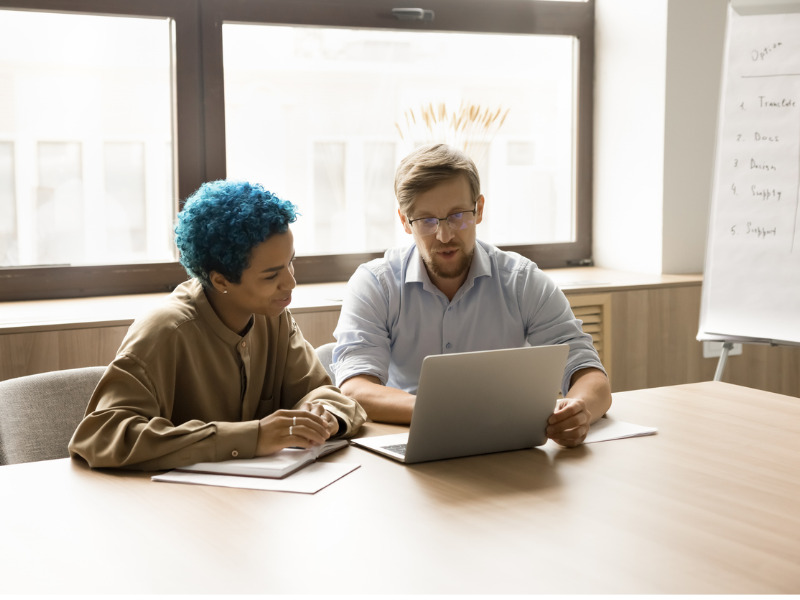 Young multiethnic business colleagues sitting at workplace, looking at laptop display, talking.