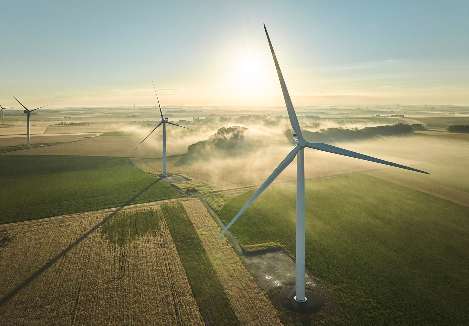 A wind turbine stands tall in an open field. Low clouds gather in the distance, amongst other wind turbines, adding a sense of depth and tranquility to the scene.