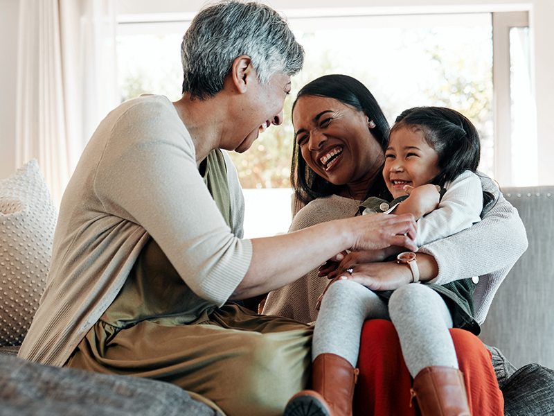 Grandmother tickling her granddaughter, who is in her mother’s arms on the couch.